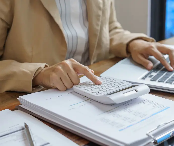 Professional in a beige blazer typing on a laptop and using a white desktop calculator over printed tax documents.